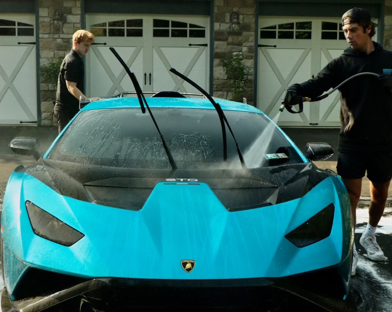 Two detailers are washing a bright cyan blue Lamborghini Huracán STO in a driveway in front of a stone garage. One man on the right uses a pressure washer to rinse the hood and windshield, while the other man on the left works near the rear of the car. The wipers are raised, and soap suds are visible on the pavement and the car's surface.