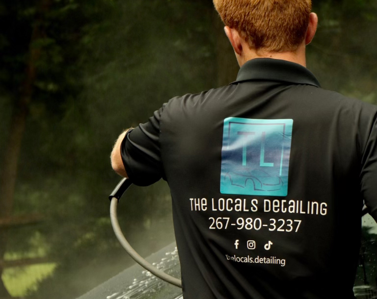 Gemini said A man with reddish-blonde hair stands with his back to the camera, wearing a black polo shirt with the logo for "The Locals Detailing." The logo features a blue square with the initials "TL" and a car outline, followed by their phone number (267-980-3237) and social media handles. He is holding a pressure washer hose and appears to be cleaning a vehicle, with a misty spray visible in the background.