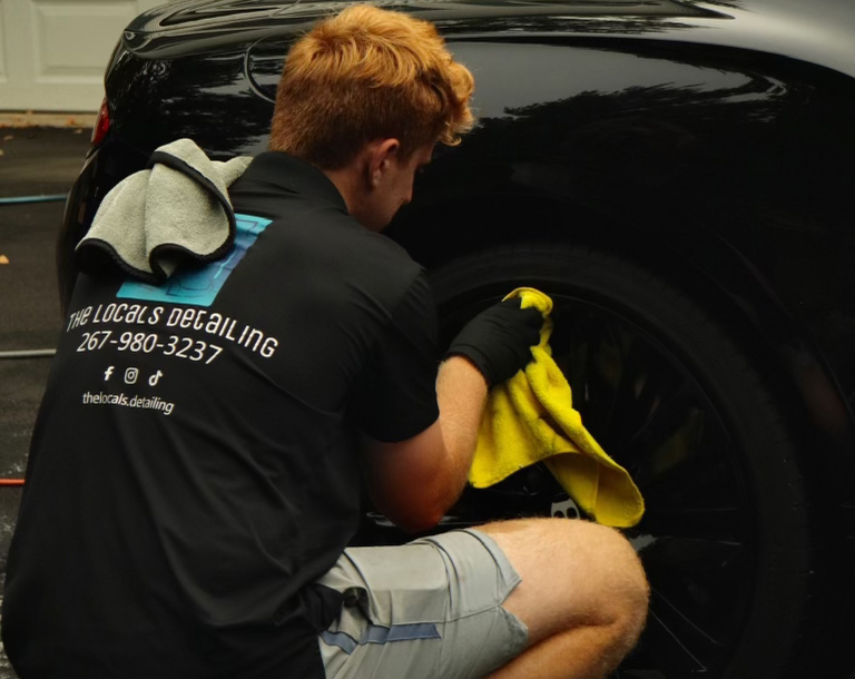 A detailer with reddish-blonde hair, wearing a black "The Locals Detailing" polo shirt, kneels to clean the black rim of a luxury vehicle. He is using a bright yellow microfiber cloth and wearing black protective gloves. A grey microfiber towel is draped over his shoulder, and the company’s contact information and social media handles are clearly visible on the back of his shirt.