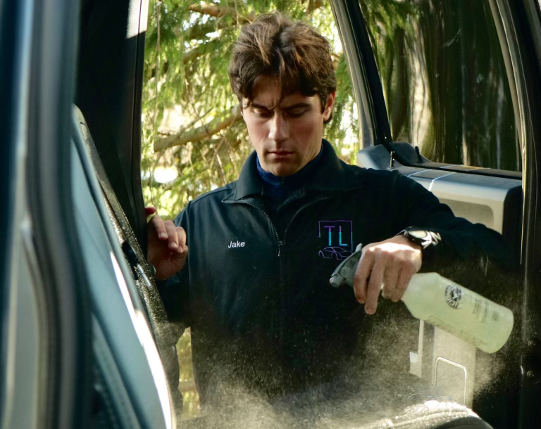 A man named Jake, wearing a black zip-up jacket with "The Locals Detailing" logo, is pictured cleaning the interior of a vehicle. He is focused on spraying a cleaning solution from a spray bottle onto the car seat, creating a fine mist in the air. The shot is framed from outside the open car door, with sunlight filtering through trees in the background.